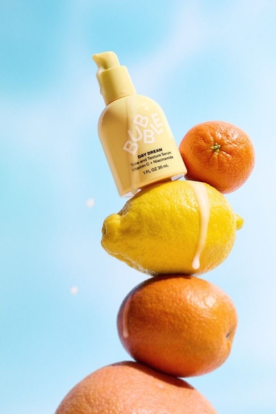 A high-key studio shot of a tone and texture serum bottle balanced on a vertical stack of citrus fruits against a bright blue sky background.